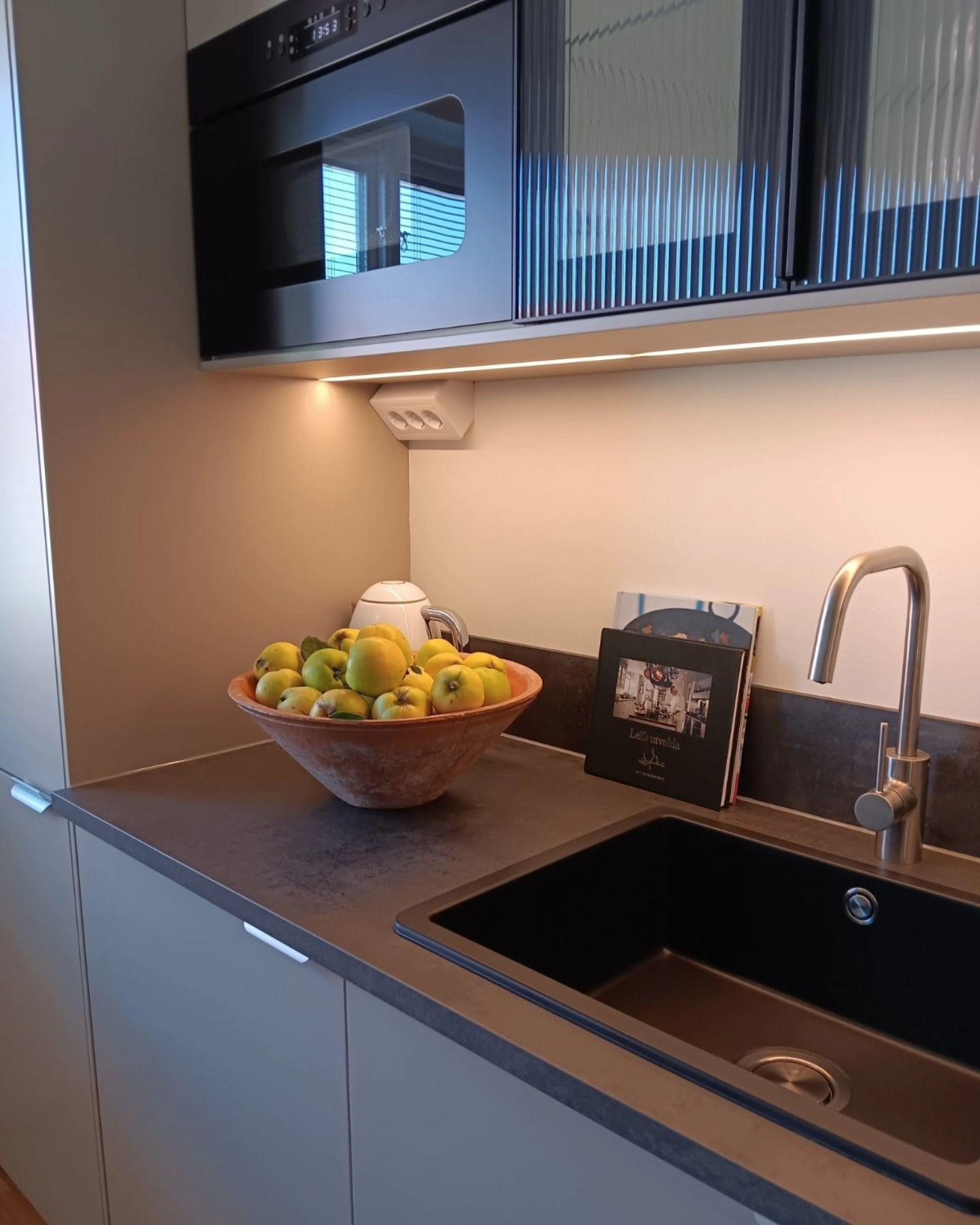 Närbild på bänkskiva med svart diskho, blandare och fruktskål under belyst väggskåp